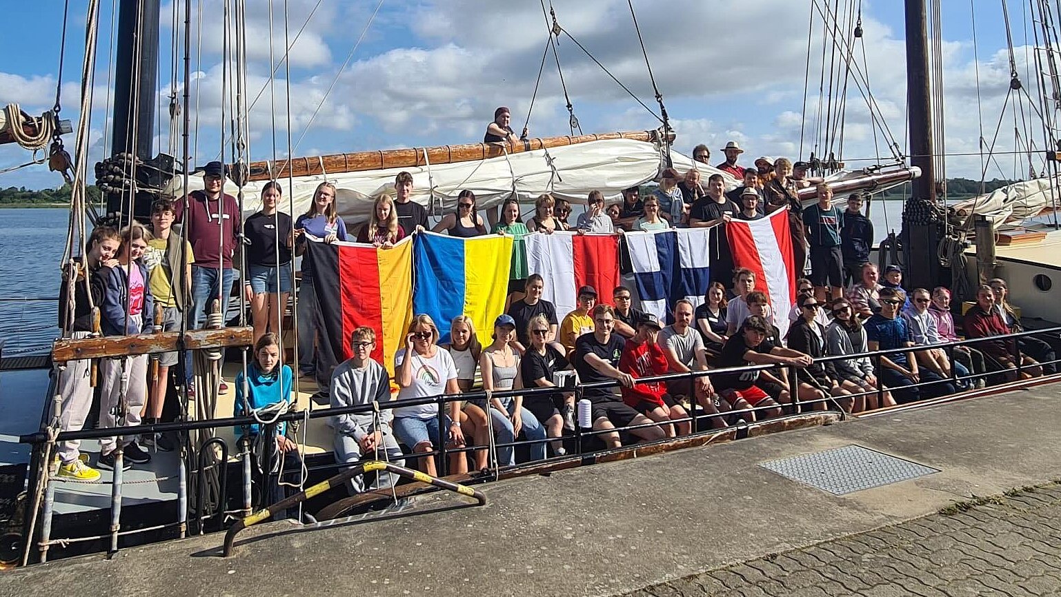 Gruppenfoto, ca. 60 Jugendliche stehen mit den Flaggen von Österreich, Finnland, Polen, Ukraine und Deutschland an Bord eines Segelschiffes.