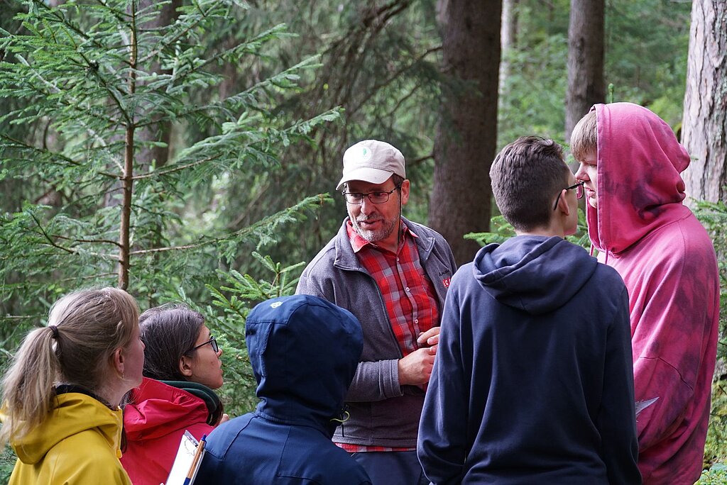 Es sind sechs Personen abgebildet, die sich im Wald befinden. Es sieht so aus, als würde eine Person den anderen etwas erklären.