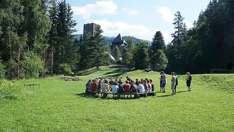 Auf einer grünen Wiese sitzen junge Menschen im Kreis, die Wiese ist von Bäumen links und rechts umgeben, im Hintergrund sind Berge und eine Burg erkennbar.