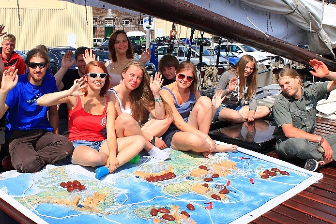 Eine Gruppe junger Menschen sitzt an Bord eines Schiffes auf einer großen Weltkarte und winkt fröhlich.