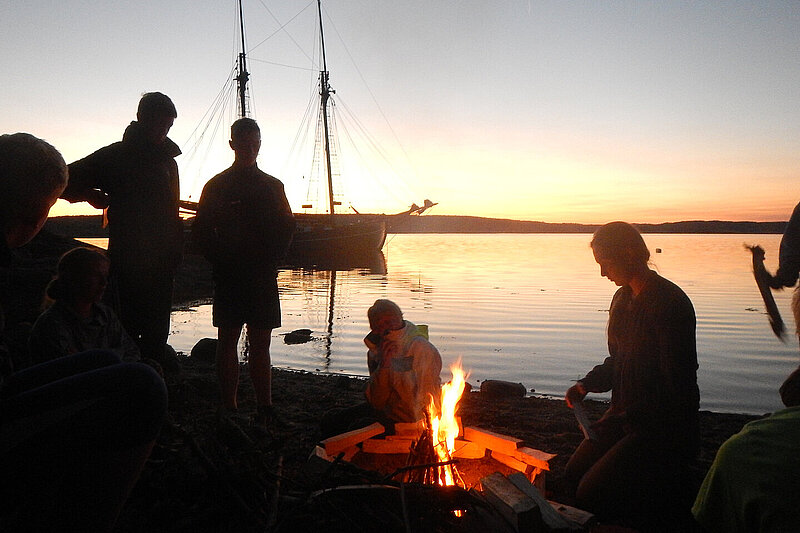 Im Vordergrund mehrere Silhouetten von Personen um ein Lagerfeuer, im Hintergrund ein Segelschiff auf dem Wasser im Sonnenuntergang.