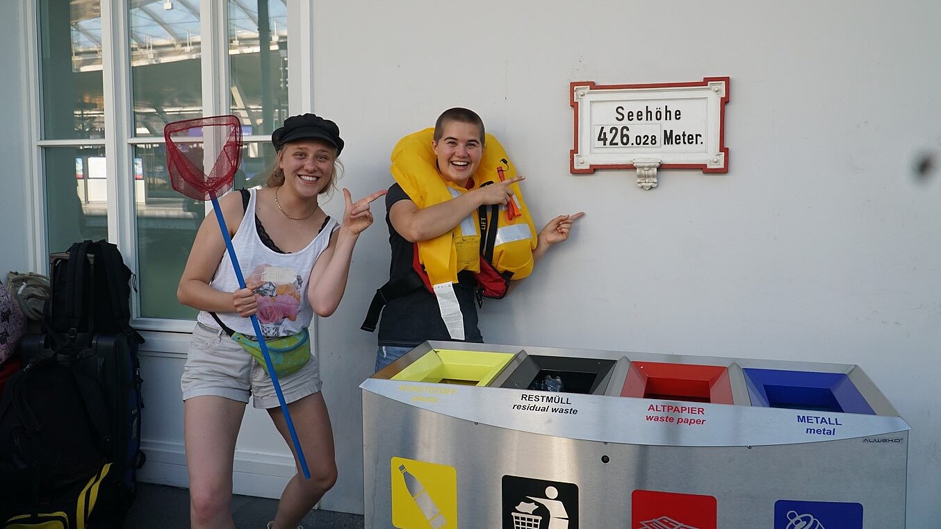 Zwei junge Menschen, eine hält einen Kescher, die andere trägt eine Schwimmweste, stehen an einem Bahnhof und deuten lachend auf ein Schild mit der Aufschrift: "Seehöhe 426,028 Meter."