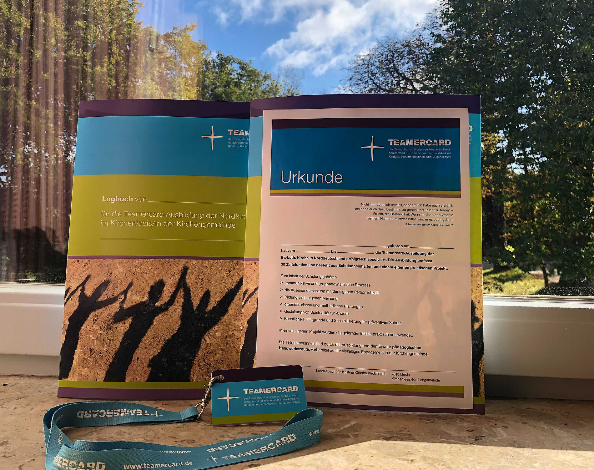 Vor einem Fenster ist die Teamercard-Urkunde, das Teamercard-Logbuch, die Teamercard- Karte sowie das Schlüsselband zu sehen 
