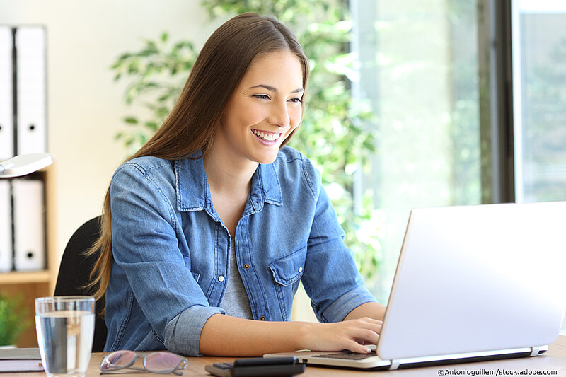 Eine junge Frau am Tisch sitzend, schreibt mit freudigem Gesichtsausdruck auf ihrem Laptop.